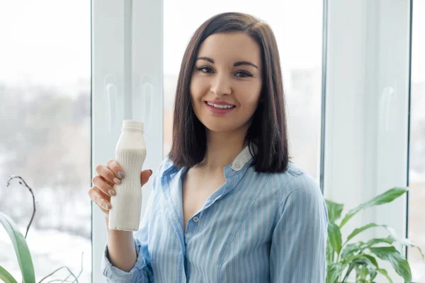Retrato matutino de una joven sonriente bebiendo leche bebiendo yogur de botella, mujer de pie en casa con camisa cerca de la ventana, comiendo comida saludable —  Fotos de Stock