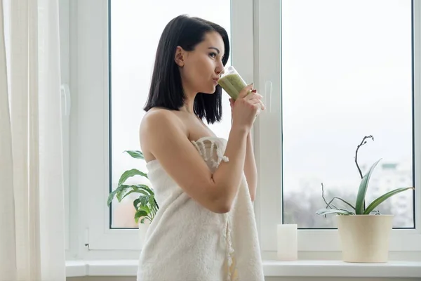 Retrato matutino de jovem mulher bonita em toalha de banho em casa perto da janela bebendo smoothie kiwi verde recém-misturado em vidro, bebida vitamínica na temporada de primavera de inverno, alimentação saudável — Fotografia de Stock