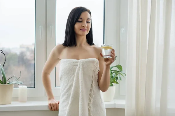 Retrato matinal de jovem mulher bonita em toalha de banho com copo de água com limão perto da janela. Estilo de vida saudável, dieta, antioxidante . — Fotografia de Stock