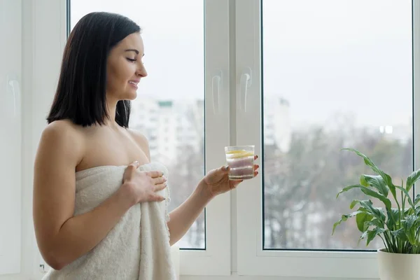 Retrato matutino de jovem mulher bonita em toalha de banho com copo de água com limão perto da janela, bebida vitamínica na estação de primavera de inverno. Estilo de vida saudável, dieta, antioxidante . — Fotografia de Stock