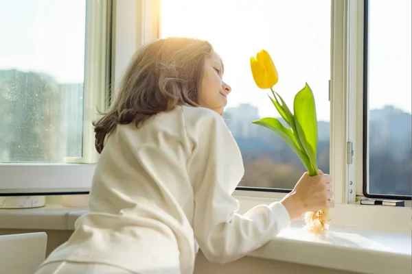 Menina criança com flor de tulipa amarela, perto do peitoril da janela olha para fora do vidro de abertura da janela, dia ensolarado da primavera — Fotografia de Stock