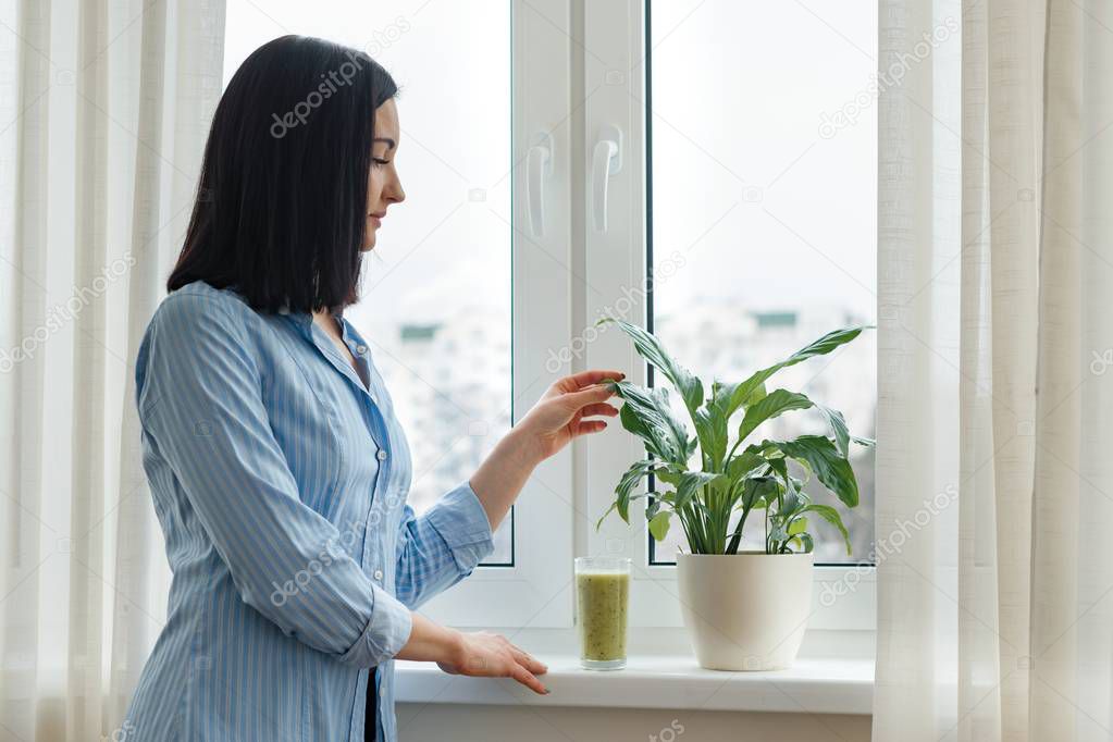 Young woman morning at home near the window drinking freshly blended green kiwi fruit smoothie in glass, vitamin drink in winter spring season, healthy food eating