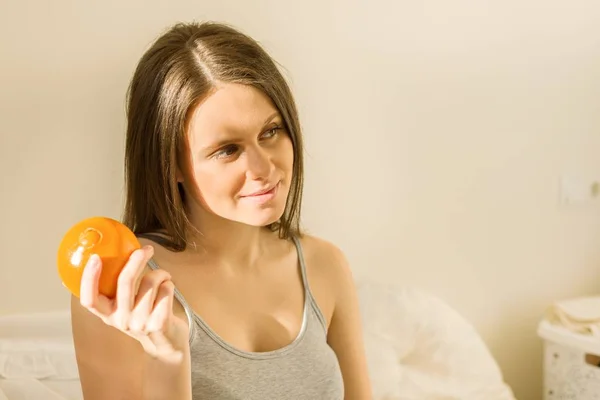 Jovem bela mulher sorridente segurando uma laranja, em casa sentado na cama — Fotografia de Stock