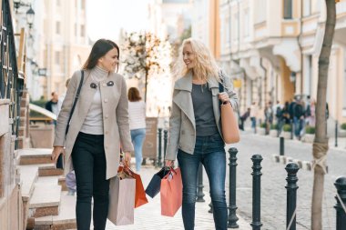 Kentsel tarzı, iki genç gülümseyen moda kadın alışveriş çantaları, güneşli sonbahar günü, altın saat ile bir şehir sokak boyunca yürüyüş