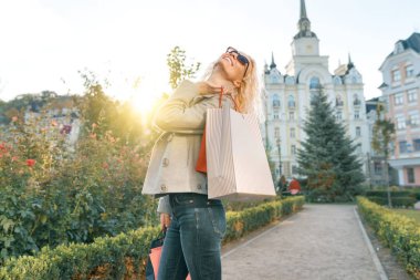 Alışveriş için çanta ile kıvırcık saçlı genç gülümseyen sarışın kadın, güneşli şehir arka plan, altın saat