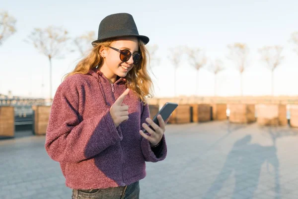 Jovem adolescente em chapéu e óculos olhando no telefone celular e mostrando indicador dedo para cima atenção, ideia, eureka, hora de ouro . — Fotografia de Stock