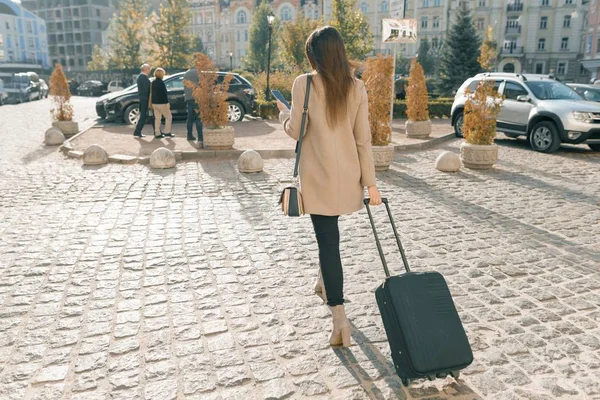 Mujer hermosa joven caminando por la calle de la ciudad con maleta de viaje y teléfono celular. Chica morena de moda, vista desde atrás, espacio para copiar — Foto de Stock