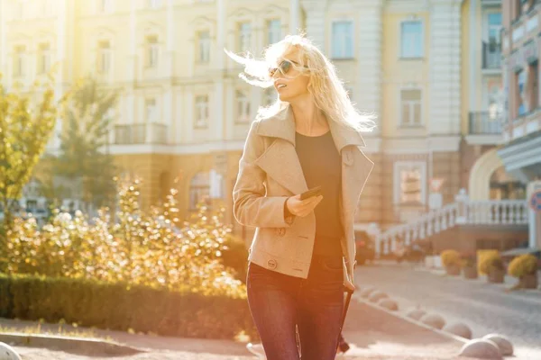 Молодая улыбающаяся блондинка с дорожной сумкой и телефоном, идущая по городской улице, женщина в солнечных очках с длинными вьющимися волосами — стоковое фото