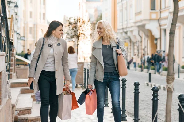 Городской стиль, две молодые улыбающиеся модные женщины, идущие по городской улице с сумками для покупок, солнечный осенний день, золотой час — стоковое фото