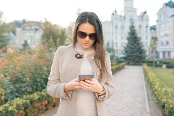 Молодая женщина пишет смс по смартфону, гуляя по солнечной улице, девушка в теплом пальто — стоковое фото