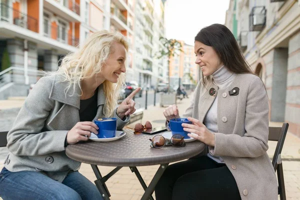 Duas jovens mulheres sorridentes em um café ao ar livre, bebendo café, conversando, rindo. Fundo de outono urbano — Fotografia de Stock