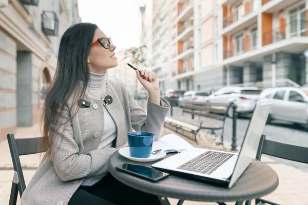 Jovem estudante bonita usando óculos casaco quente sentado em um café ao ar livre com laptop copo de computador de bebida quente segurando caneta em suas mãos escrevendo em notebook, cidade rua fundo — Fotografia de Stock