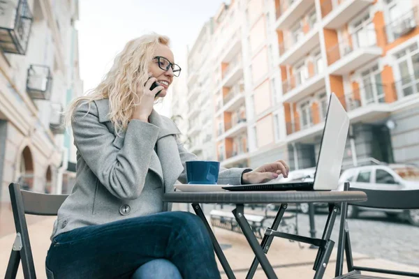 Giovane bella donna d'affari bionda in occhiali in abiti caldi seduto in un caffè all'aperto con computer portatile bere tazza di caffè e parlare al telefono, sfondo strada della città — Foto Stock