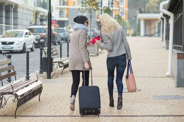 Dwie młode uśmiechnięte kobiety z walizki spacerowej wzdłuż ulicy miasta. Tło miejskie, jesienny dzień, widok z tyłu — Zdjęcie stockowe