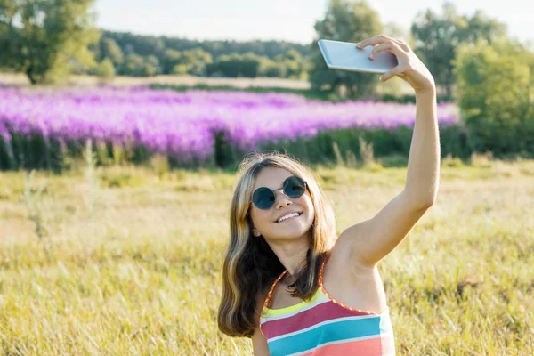 Jovencita adolescente caminando en la naturaleza es fotografiado en el teléfono inteligente móvil, soleado verano día fondo púrpura lavanda campo — Foto de Stock