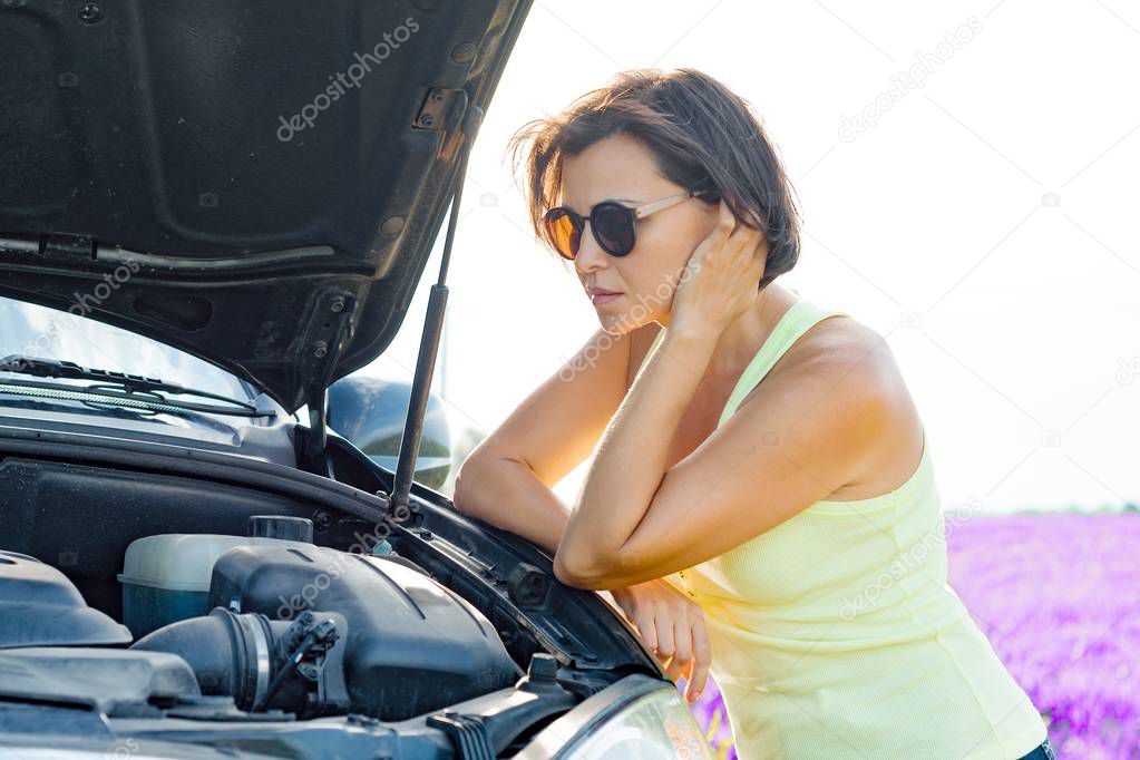 Distressed woman driver standing near broken car, raised hood. Summer nature background, lavender field
