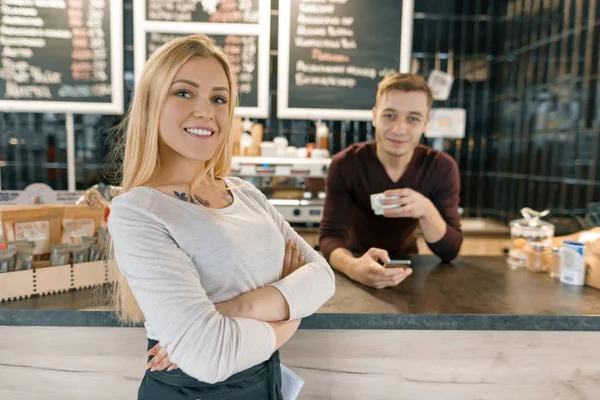 Joven pareja sonriente de trabajadores de la cafetería, hombre y mujer posando cerca del mostrador del bar y la máquina de café con taza de café fresco. Trabajo en equipo, personal, pequeñas empresas, concepto de personas —  Fotos de Stock