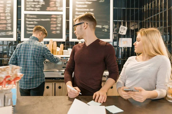 Ungdomarna arbetar i kafé, man och kvinna nära bar counter, ta emot order via telefon, barista på bakgrunden av kaffebryggare. Lagarbete, personalen, småföretag, människor-konceptet — Stockfoto