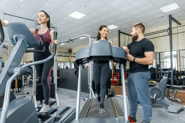 Jóvenes mujeres fitness sonrientes con entrenador personal un hombre atlético adulto en la cinta de correr en el gimnasio. Deporte, trabajo en equipo, entrenamiento, concepto de estilo de vida saludable — Foto de Stock