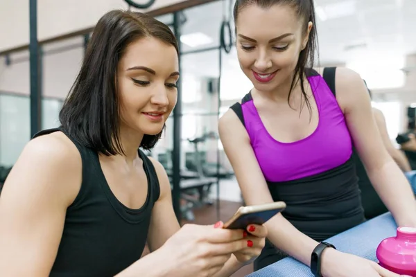 Zwei junge Sportlerinnen im Fitnessstudio unterhalten sich lächelnd mit Fitnessmatten und schauen aufs Handy. Training, Teamwork, gesundes Lebensstilkonzept. — Stockfoto