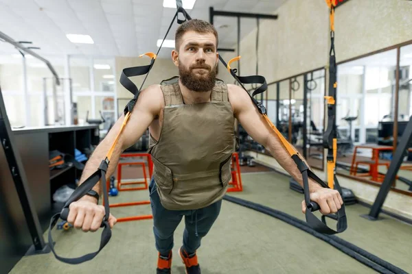 Muscular bearded man dressed in military weighted armored vest doing exercises using straps systems in the gym. Sport, training, bodybuilding and healthy lifestyle concept.