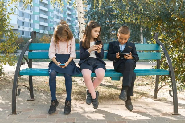 Grupo de niños de 7, 8 años con teléfonos móviles, mirando con entusiasmo en los teléfonos inteligentes, escolares con mochilas, fondo al aire libre. Educación, amistad, tecnología y concepto de personas —  Fotos de Stock