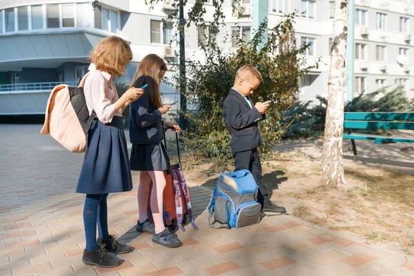 Niños de edad elemental con teléfonos inteligentes, mochilas, fondo exterior. Educación, amistad, tecnología y concepto de personas —  Fotos de Stock