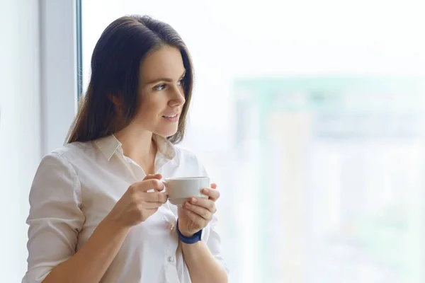 Jeune femme en chemise blanche avec une tasse de café près de la fenêtre, femme brune regarde par la fenêtre en souriant — Photo