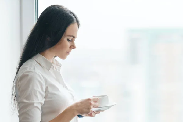 Retrato de jovem empresária em camisa branca com xícara de café, mulher sorridente desfrutando de seu café aromático matutino, escritório em arranha-céu dia nublado, espaço de cópia — Fotografia de Stock