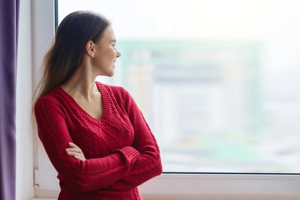 Muda tersenyum percaya diri wanita berdiri di dekat jendela, perempuan menyeberang lengannya, wanita dalam sweter rajutan — Stok Foto