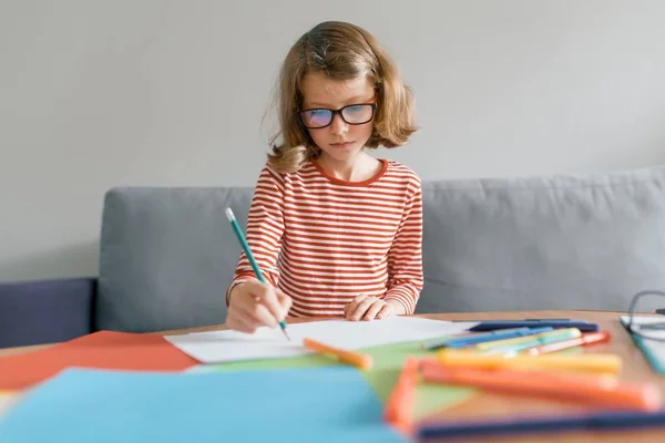 Chica de 8 años sentada en sofá en casa dibujando escritura con lápiz en cuaderno. Rubia infantil con gafas estudiando en casa — Foto de Stock