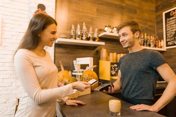 Menina cliente cafetaria paga café com cartão de crédito, barista segurando uma máquina leitor de cartão de crédito — Fotografia de Stock