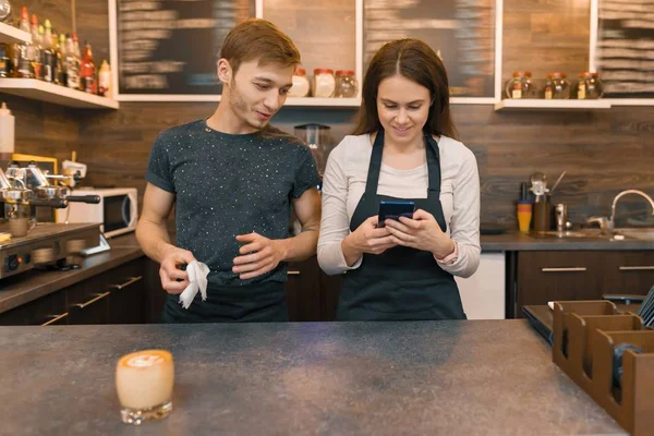 Młodzi pracownicy kawiarni mężczyzna i kobieta za barem licznika, rozmowa, patrząc na smartphone — Zdjęcie stockowe