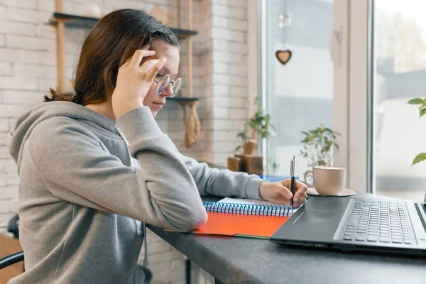 Ritratto di giovane studentessa, studentessa di liceo in caffetteria con computer portatile e tazza di caffè, ragazza sta studiando, scrivendo nel quaderno — Foto Stock
