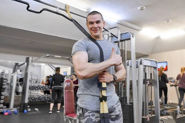 Ritratto di giovane uomo sorridente, muscoloso e giovane in palestra. Maschio con cinghie da fitness loop — Foto Stock