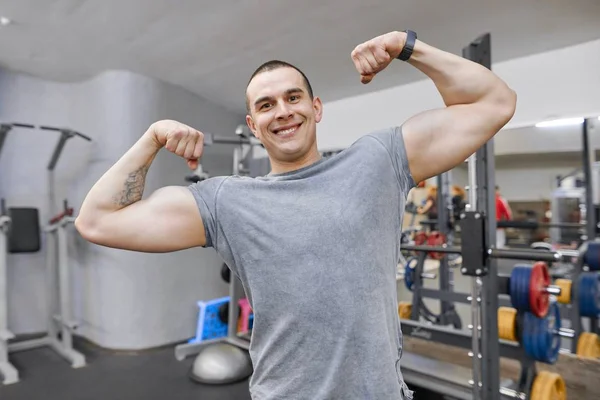 Νέοι ισχυρή χαμογελώντας μυώδης άνδρας σε γυμναστήριο δείχνει έντονη μυϊκή αγκαλιά. — Φωτογραφία Αρχείου