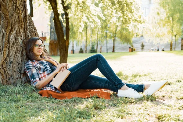 Krásná žena s brýlemi čtení knihy sedí na trávě vedle stromu v parku při západu slunce. — Stock fotografie