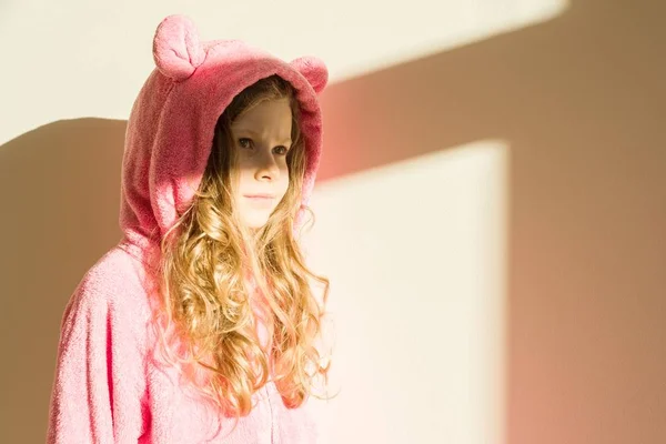 Retrato de uma menina em pijama rosa quente macio. Menina de 7 anos, loira com cabelos longos encaracolados, em um capuz, olhando para longe. Fundo parede da casa de luz, iluminação solar da janela, espaço de cópia — Fotografia de Stock