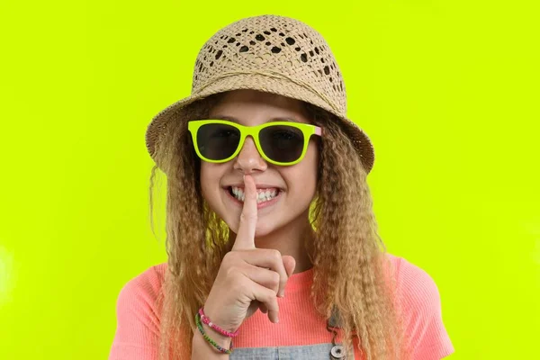 Retrato de menina adolescente em óculos de sol e chapéu de palha com um indicador perto dos lábios, mostrando sinal de silêncio, gesto secreto, fundo estúdio amarelo — Fotografia de Stock