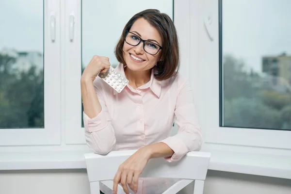 Портрет улыбающейся женщины в очках и легкой рубашке с волдырем таблеток, держащей и показывающей таблетки у окна — стоковое фото
