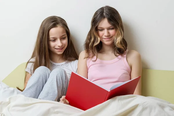 Dos niñas hermanas sentadas en casa en la cama con un cuaderno escolar leyendo y estudiando preparándose para la escuela en casa. Comunicación y aprendizaje —  Fotos de Stock