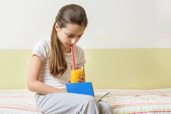 Девочка сидит дома в постели со стаканом свежего апельсинового сока и читает книгу — стоковое фото