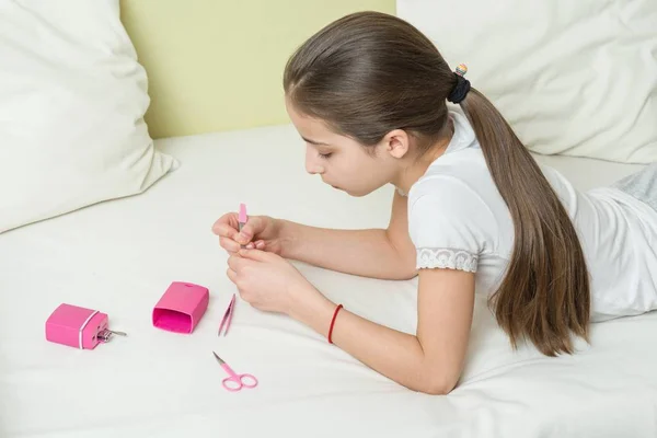 Het meisje is 10 jaar oude thuis op het bed in haar eigen kleren, nails haar nagels met behulp van manicure accessoires — Stockfoto