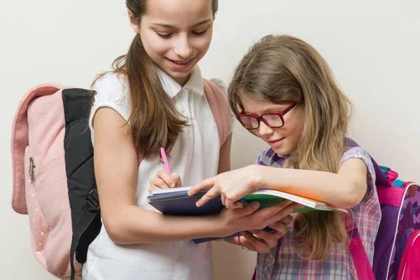 Dos chicas sonrientes con bolsos escolares. Colegialas de 7 y 10 años hablando en la escuela. Niña escribe en un cuaderno . — Foto de Stock