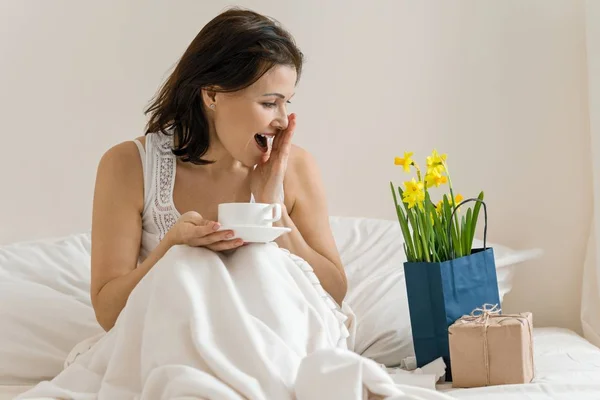 Mellersta år kvinna är nöjd med gåva, blombukett, sitter i morgon i sängen med kopp kaffe. Känslor av lycka, glädje, överraskning — Stockfoto