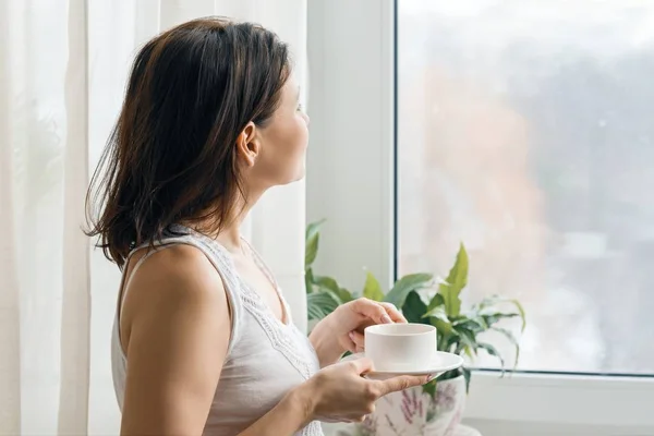 Copa de café fresco de la mañana en las manos de la mujer de pie y mirando por la ventana — Foto de Stock