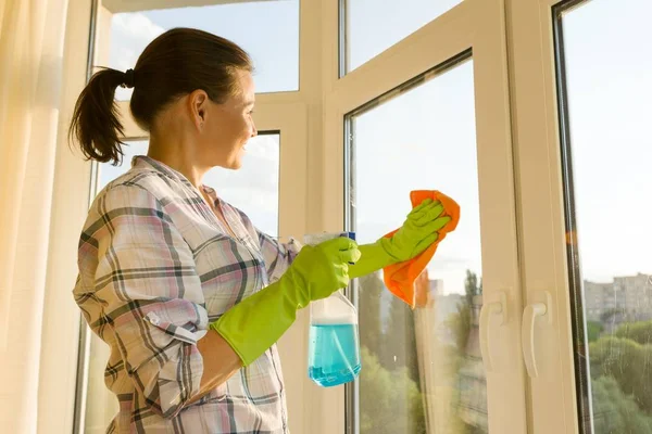 Mujer madura en casa lavando la ventana con guantes protectores de goma con detergente pulverizador y trapo de microfibra . —  Fotos de Stock