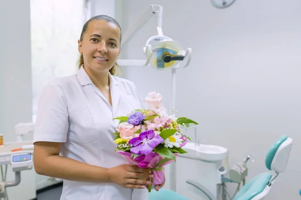 Vrouwelijk tandarts glimlachend, met een boeket van bloemen, in tandheelkundige kantoor. Dag van de nationale tandarts. — Stockfoto