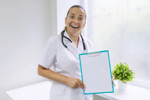 Adulto sorridente mulher médico com uma folha em branco na área de transferência para o seu texto, janela do hospital de fundo — Fotografia de Stock