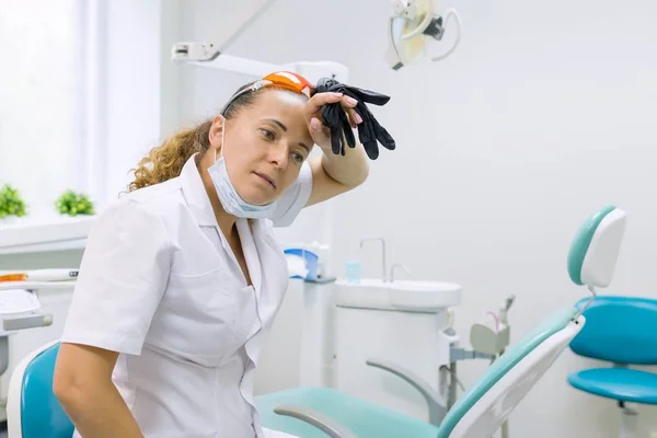 Vermoeide vrouw tandarts, achtergrond tandheelkundige bureaustoel. Vrouwelijke tandarts opstijgen handschoenen vegen haar voorhoofd — Stockfoto